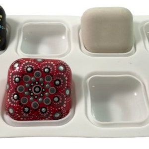 May include: White silicone mold with eight square cavities. Three of the cavities are filled with square-shaped painted rocks. One rock is black with white and gold dots, one is red with silver and black dots, and one is purple with gold and white dots.