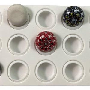 May include: A white silicone mold with 12 round cavities for making small, round, decorative rocks. The mold is shown with three painted rocks, one gray, one red with white dots, and one black with gold and white dots.