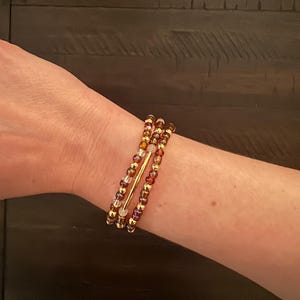 May include: A set of three beaded bracelets in shades of gold, amber, and purple. The bracelets feature small, round beads and a gold-colored bar. The bracelets are stacked on a wrist.
