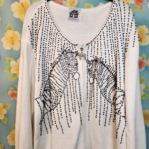 May include: A white long-sleeve zip-up cardigan featuring black beaded vertical lines and two black beaded zebra motifs on the front. The zipper pull is shaped like a small zebra. A Storybook Knits label is sewn inside the V-neck collar.