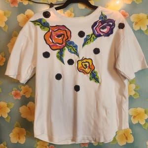 May include: White t-shirt with embroidered floral design. The shirt features three colorful roses in orange, purple, and yellow, with green and blue leaves. Black polka dots are scattered across the shirt.