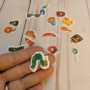May include: A set of colorful stickers featuring various foods and objects from the children's book The Very Hungry Caterpillar. The stickers include a green caterpillar, a red apple, a green pear, a yellow cupcake, a slice of watermelon, a piece of cheese, a lollipop, a strawberry, a piece of chocolate cake, a butterfly, a plum, a sausage, and a green leaf.