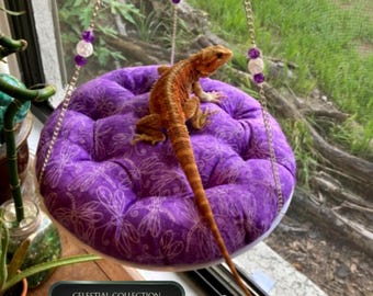 May include: A round purple tufted cushion with a white dragonfly pattern, suspended by silver chains adorned with purple and clear beads. A bearded dragon rests on the cushion. The text CELESTIAL COLLECTION is visible in a dark rectangular label.