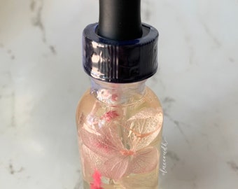 May include: A clear glass bottle with a black dropper top filled with a light yellow liquid and a single white flower with pink accents.