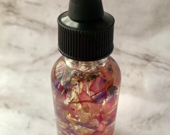 May include: A clear glass bottle with a black dropper top filled with a light yellow oil infused with dried pink, purple, and white flowers.