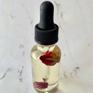 May include: A clear glass bottle with a black dropper top filled with a light yellow liquid and dried rose petals.