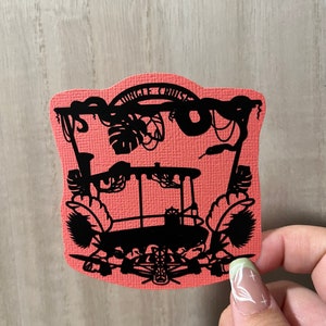 May include: A coral-colored paper cutout featuring a black silhouette of a Jungle Cruise scene. The design includes a boat, jungle foliage, and the words "Jungle Cruise" at the top. The cutout is held by a hand with a light green and white nail design.
