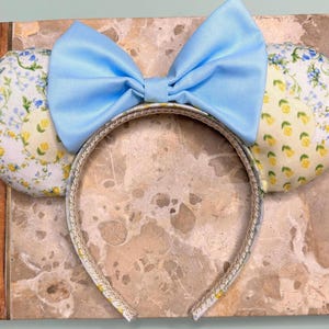 May include: Light blue bow headband with floral print ears. The ears feature a white background with yellow lemons and blue flowers.