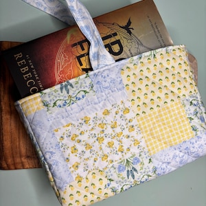 May include: A quilted book bag with a patchwork design featuring yellow and blue floral patterns. The bag has a blue and white ribbon handle and a book titled "Flames" is inside.