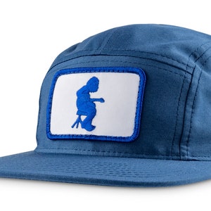 Peut inclure: Une casquette bleue à 5 panneaux avec un patch blanc représentant une silhouette bleue d'une personne assise sur un tabouret.
