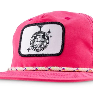Peut inclure: Un chapeau de camionneur rose avec un patch blanc représentant un ballon disco noir et blanc suspendu au plafond. Le chapeau a une bordure en corde blanche et multicolore autour du bord.