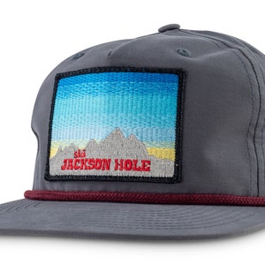 Puede incluir: Una gorra de béisbol gris con ribete de cuerda roja y un parche con una escena de montaña y el texto "ski JACKSON HOLE" en rojo.