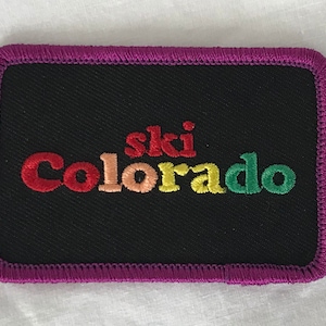 May include: A black embroidered patch with a purple border. The text "ski Colorado" is embroidered in rainbow colors.