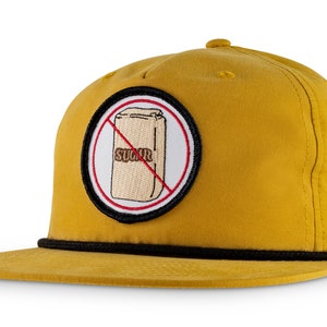 Peut inclure: Une casquette de baseball jaune avec une visière noire et un patch noir et blanc qui dit "No Sugar" avec une ligne rouge traversant un sac de sucre.