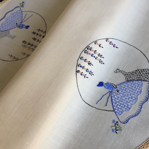 May include: A white linen tablecloth with a scalloped edge and embroidered circles featuring blue and white figures in a garden setting.