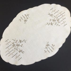 May include: Oval, cream-coloured embroidered doily with a scalloped edge. The doily features embroidered floral and geometric designs in a muted brown colour. A decorative piece for a table or dresser.