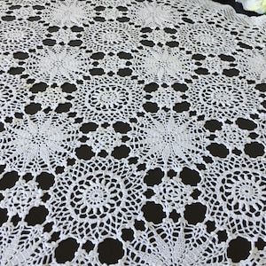 May include: A white crocheted tablecloth featuring a repeating pattern of circular motifs connected by intricate openwork stitching. The design consists of large, star-like floral shapes alternating with smaller circular elements, creating a lace-like texture.