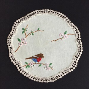 Può includere: Sottobicchiere rotondo ricamato color crema con un uccello posato su un ramo fiorito. L'uccello presenta dettagli marroni, rossi e blu. Il sottobicchiere ha un bordo smerlato con un bordo in pizzo color crema. Il ricamo è dettagliato.