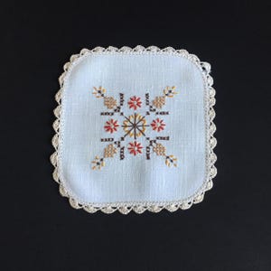 May include: A square, light blue embroidered coaster with a cream-colored, scalloped lace edge. The center features a cross-stitch design in red, brown, and yellow floral patterns. The coaster is likely handmade.