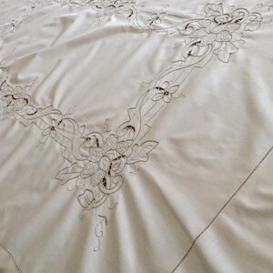 May include: Cream-coloured tablecloth with detailed floral embroidery. The embroidered design features stylized flowers and leaves, creating a delicate border. The fabric has a subtle sheen.