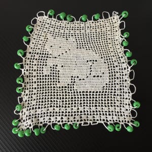 Peut inclure: Un napperon carré en crochet blanc cassé présentant un motif central de silhouette de chat créé avec un point en forme de grille. Les bords du napperon sont finis avec une série de petites perles vert vif attachées aux boucles de la bordure.