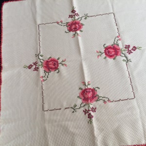Puede incluir: Mantel blanco con un dise&ntilde;o floral bordado en rojo y verde. El dise&ntilde;o presenta cuatro rosas en las esquinas de un cuadrado.
