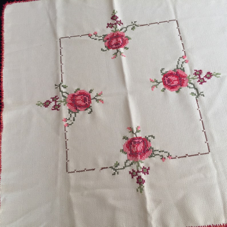 Puede incluir: Mantel blanco con un dise&ntilde;o floral bordado en rojo y verde. El dise&ntilde;o presenta cuatro rosas en las esquinas de un cuadrado.