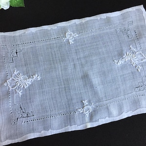 May include: A rectangular white linen doily featuring intricate white floral embroidery and delicate cutwork borders. The piece displays detailed needlework patterns in the corners and along the edges, showcasing a fine, semi-sheer fabric texture.