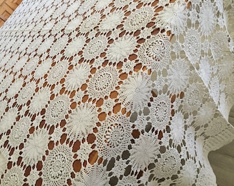 May include: A white crocheted tablecloth with a delicate floral pattern. The tablecloth is made of a fine, open weave and has a scalloped edge.