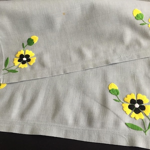 May include: Two grey linen napkins with yellow and black embroidered flowers. The flowers have a simple design with a yellow centre and black centre.