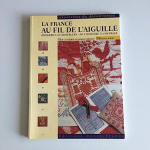 May include: A book titled "LA FRANCE AU FIL DE L'AIGUILLE" with French text and images of embroidery and lace. The cover is cream-colored with red and white illustrations, including a rooster and various needlework samples.