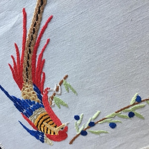 May include: White embroidered fabric with a colourful bird design. The bird has red, blue, yellow, and brown feathers. A branch with green leaves and blue berries is also embroidered on the fabric.