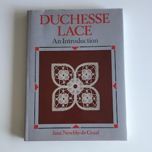 May include: A book titled "DUCHESSE LACE An Introduction" by Jane Newble-de Graaf. The cover is gray with red text and accents. The center features a white lace design on a maroon background.