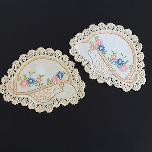 May include: Two embroidered coasters with a basket design. Each coaster features a light blue background with a basket outline in brown and a floral arrangement in pink, blue, and orange. The edges are trimmed with a cream-colored lace.