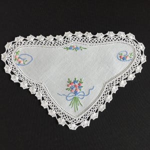 Pode incluir: Um centro de mesa triangular de tecido branco com delicados ramos de flores bordados à mão em rosa, azul e verde. A peça é finalizada com uma intrincada borda de renda de crochê branca que segue a forma pontiaguda do tecido.