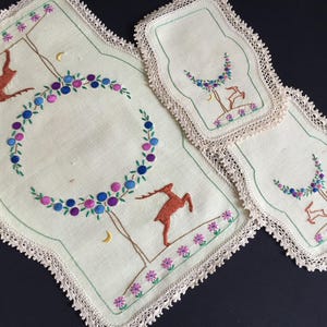 Peut inclure: Ensemble de sets de table et dessous de verre brodés en lin. Le tissu crème présente un motif de cerf et de fleurs dans les tons marron, bleu, violet et rose. Les bords sont ornés d'une dentelle délicate.