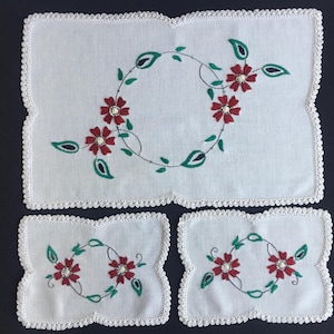 Può includere: Un set da tavola bianco ricamato e due sottobicchieri abbinati. Ogni pezzo presenta un motivo floreale con fiori rossi, foglie verdi e un bordo all'uncinetto bianco. Il set da tavola misura circa 30 x 41 cm.