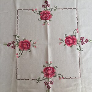 Puede incluir: Un mantel blanco con un borde bordado marr&oacute;n y cuatro rosas rojas en las esquinas.