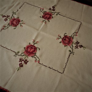 Puede incluir: Un mantel blanco con un dise&ntilde;o cuadrado bordado con rosas rojas y hojas verdes.