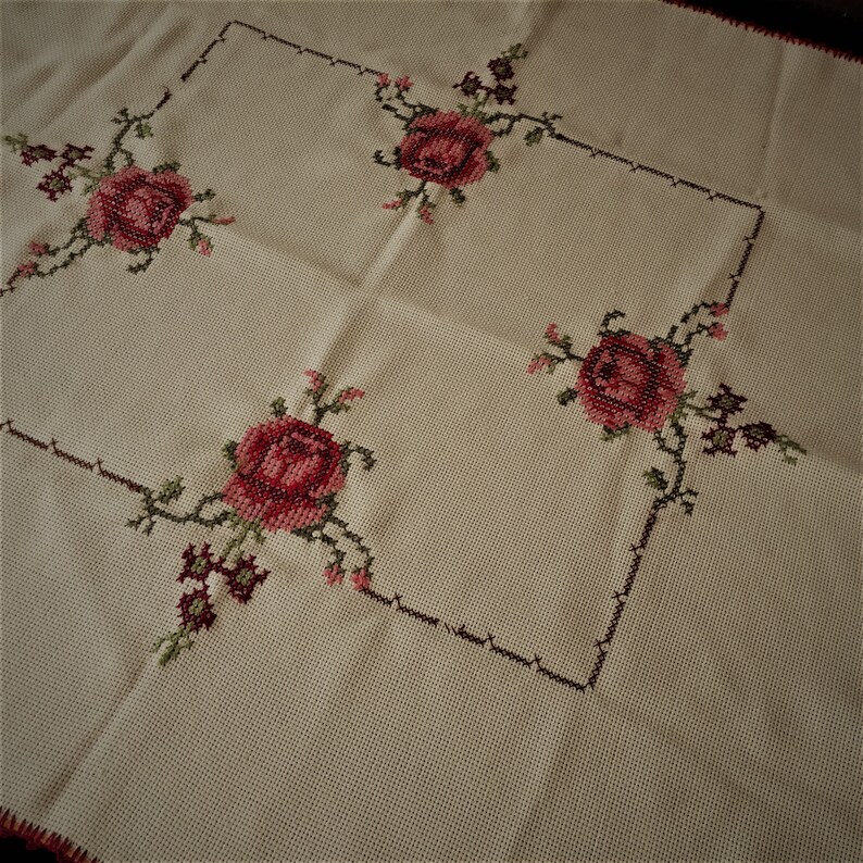 Puede incluir: Un mantel blanco con un dise&ntilde;o cuadrado bordado con rosas rojas y hojas verdes.