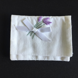 May include: A rectangular white fabric pouch featuring an embroidered design of three lavender sprigs in purple and green thread. A white satin ribbon is tied into a bow over the stems of the lavender, secured to the front of the white textile.