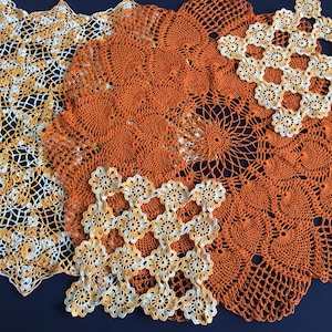 Può includere: Una collezione di centrini all'uncinetto fatti a mano in varie dimensioni e motivi. I centrini presentano disegni intricati nelle tonalità dell'arancione, crema e bianco. La disposizione mette in mostra la trama e i dettagli degli oggetti fatti a mano.