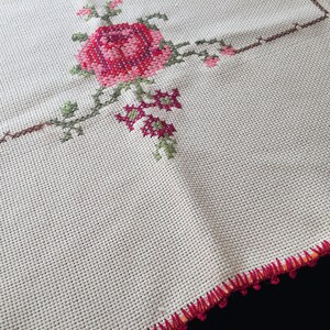 Puede incluir: Un mantel blanco con un borde de ganchillo y un dise&ntilde;o floral bordado en rosa y verde.