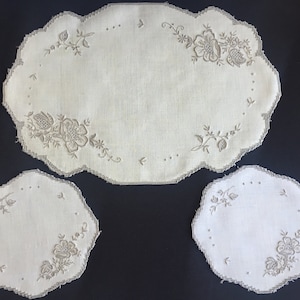 Op de afbeelding: Drie witte, geborduurde linnen placemats met een bloemenmotief. De grootste placemat is ovaal en de twee kleinere placemats zijn rond.
