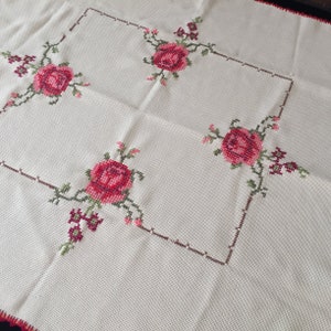Puede incluir: Un mantel blanco con un borde bordado marr&oacute;n y verde y rosas rojas en las esquinas.