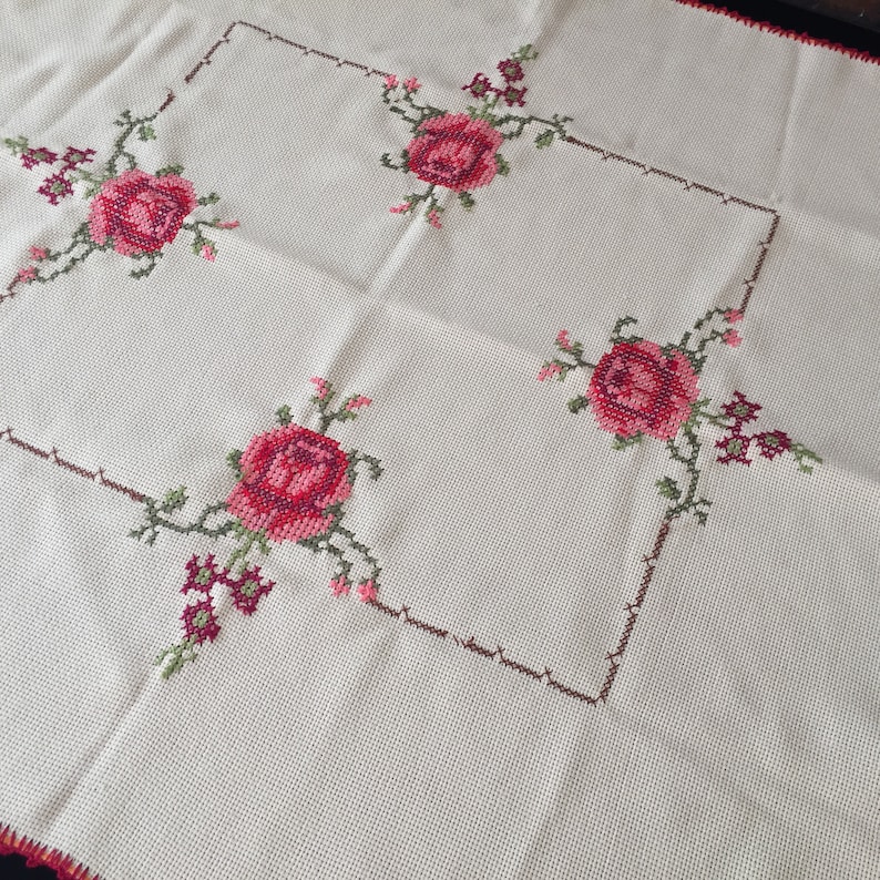 Puede incluir: Un mantel blanco con un borde bordado marr&oacute;n y verde y rosas rojas en las esquinas.