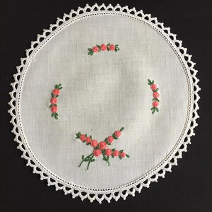 Pode incluir: Uma toalha de mesa redonda bordada branca com uma borda de renda ondulada. A toalha de mesa apresenta rosas cor-de-rosa bordadas com folhas verdes dispostas em um padrão floral. Uma peça clássica para decoração de casa.