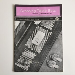 Könnte beinhalten: Schwarz-Weiß-Bild eines Vintage-Nähbuchs mit dem Titel "Dressing Table Sets" mit Häkelmustern. Das Cover ist rosa und zeigt einen Schminktisch mit Häkeldeckchen, einer kleinen Schmuckschatulle und flauschigen Hausschuhen.