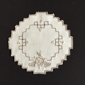 May include: A white, circular doily with an intricate embroidered floral design. The doily has a scalloped edge and geometric patterns, with delicate floral accents. The embroidery is in a neutral tone, creating a vintage aesthetic.