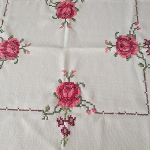 Puede incluir: Mantel blanco con un borde bordado marr&oacute;n y cuatro rosas rosas con hojas verdes.